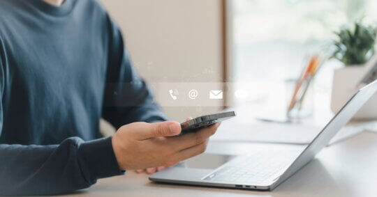 A person holding a smartphone in one hand while working on a laptop, representing managing a PeekYou opt out request online.
