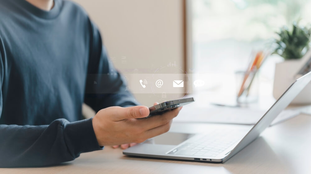 A person holding a smartphone in one hand while working on a laptop, representing managing a PeekYou opt out request online.